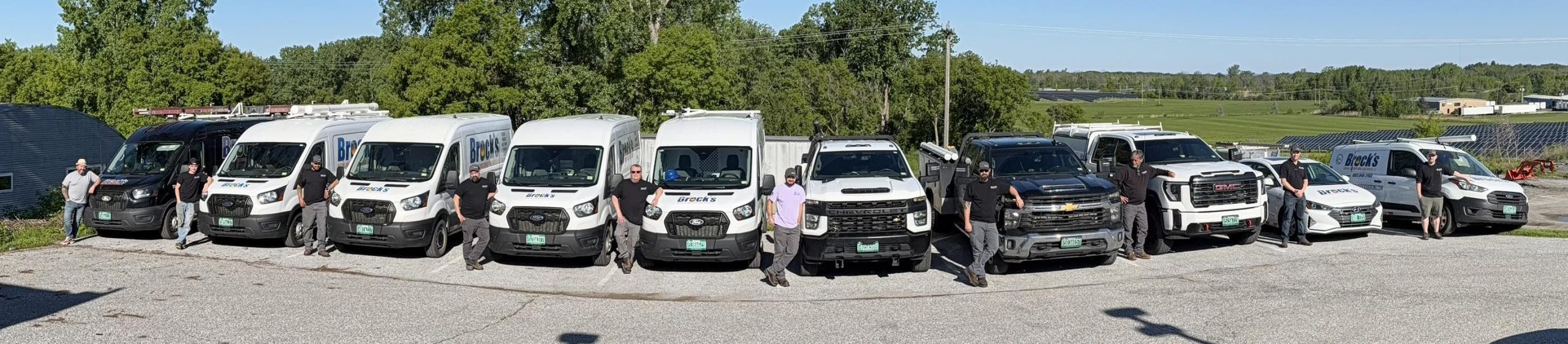 Brock's Heating service team standing in front of a fleet of company vans and trucks parked outside on a sunny day.